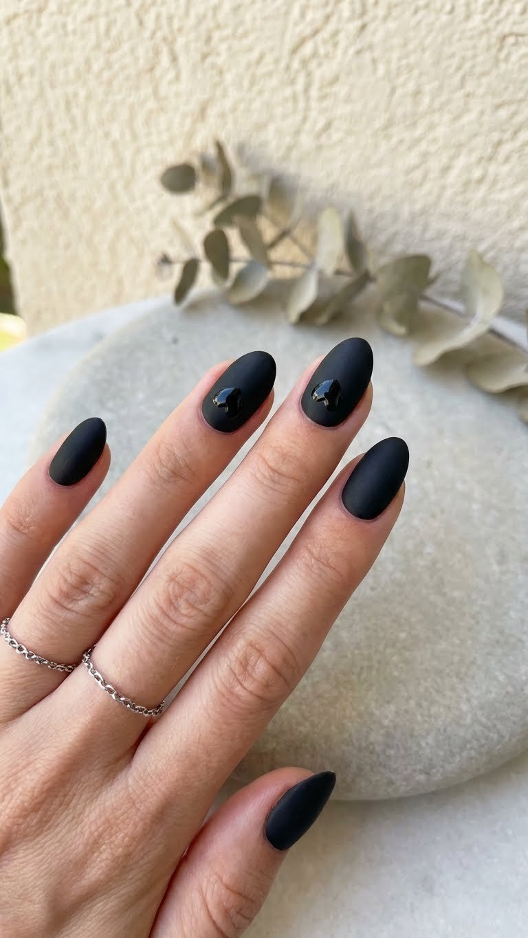 Chic minimalist goth nails: short matte black almond nails with glossy black heart details, silver ring, on a light gray surface.