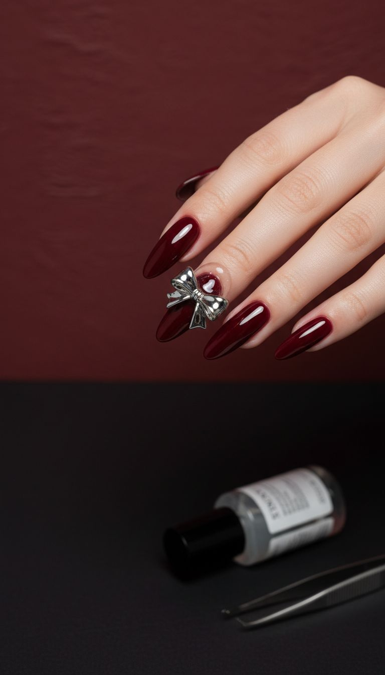 Elegant dark red almond nails with glossy silver 3D bow charm, builder gel details on black matte surface.
