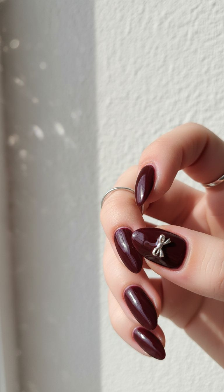 Elegant glossy burgundy almond nails, tiny intricate silver bow charm on ring finger, sleek ring, natural light.