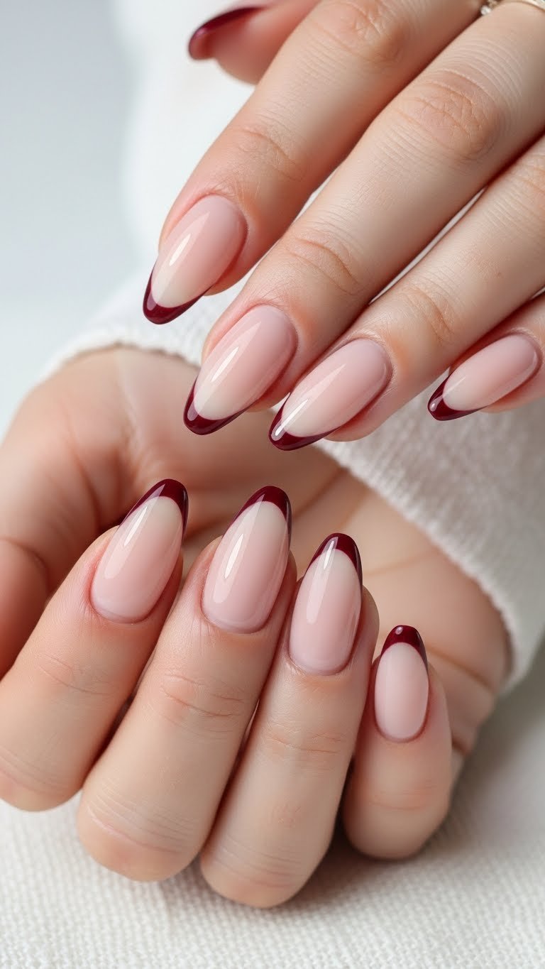 Minimalist almond nails with nude base and precise burgundy French tips on white linen, delicate jewelry.