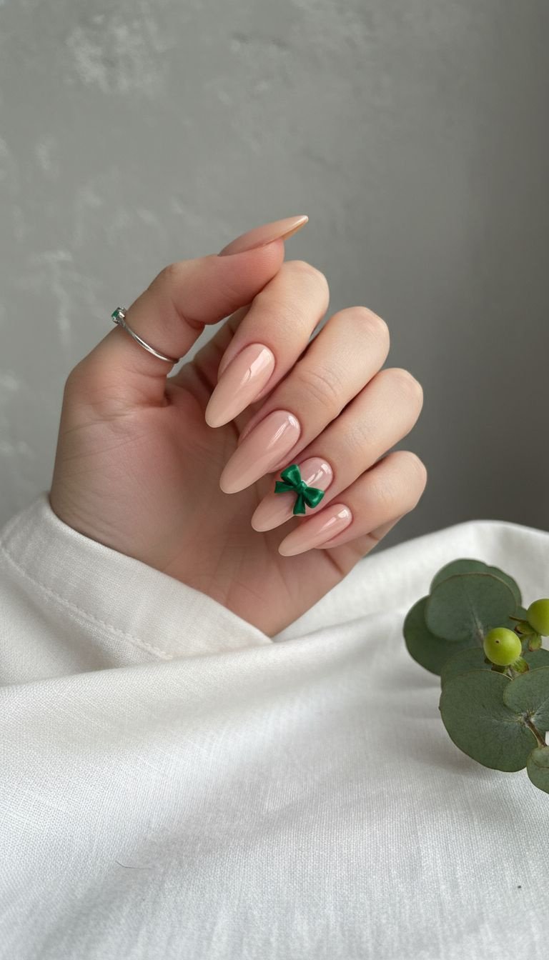 Minimalist nude acrylic nails feature a vibrant emerald green 3D bow on the ring finger. Polished design on white linen with greenery.
